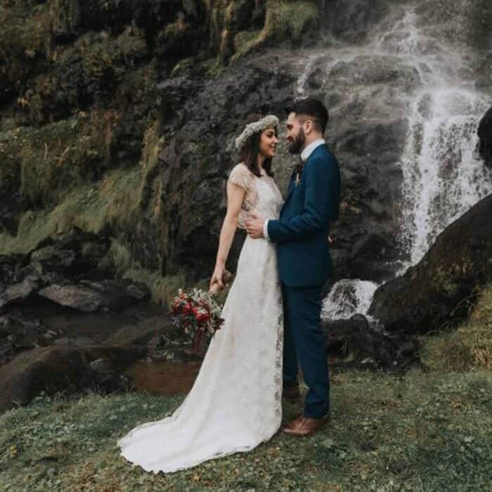 A groom in a green suit embraces his bride wearing a long white gown and a floral crown in front of a waterfall in south Iceland.