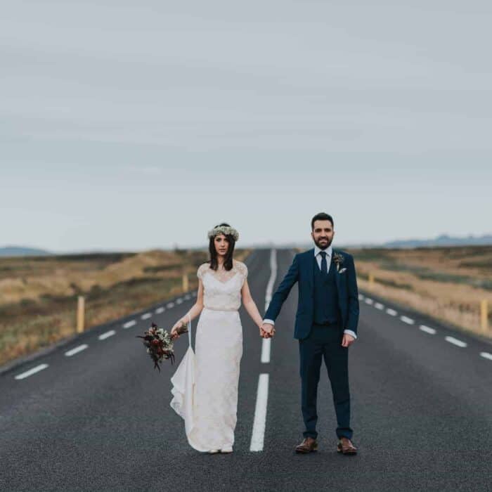 Bride in white dress and groom in suit stand in the middle of a quiet country road in south Iceland.