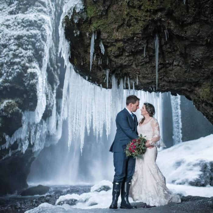 Bride in white dress and groom in suit and rain boots stand in front of an icy waterfall in south Iceland.