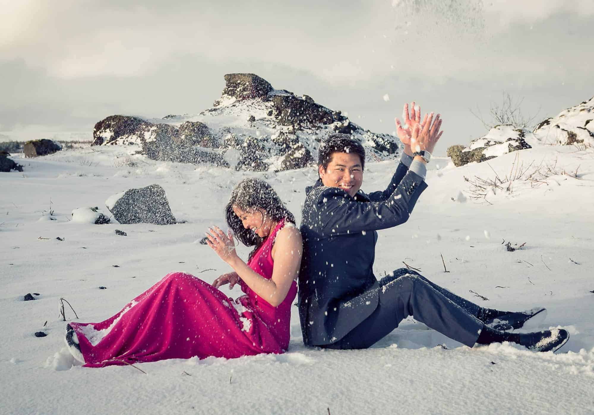 Bride wearing pink dress and groom wearing suit sit back to back on the snowy ground and toss snow overhead.