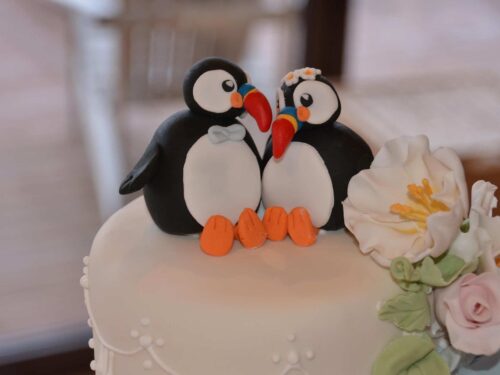Two puffins made of sugar sit atop a white wedding cake decorated with sugar flowers.