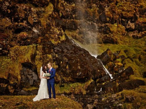 Bride wearing white dress and groom wearing blue suit embrace while standing underneath a small waterfall in south Iceland.