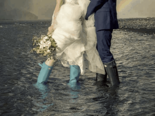 Wedding couple wearing blue boots and wedding clothes stand in the river underneath Skógafoss.
