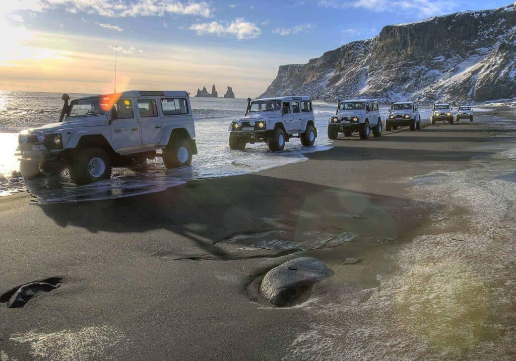 A line of white super jeeps driving across a black sand beach in the town of Vík in south Iceland.