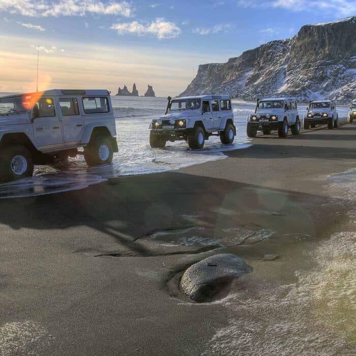 A line of white super jeeps driving across a black sand beach in the town of Vík in south Iceland.