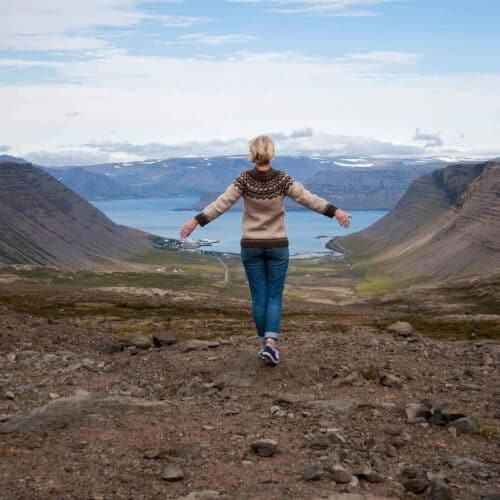 Woman wearing Icelandic lopapeysa sweater stands high above a fjord in west Iceland.