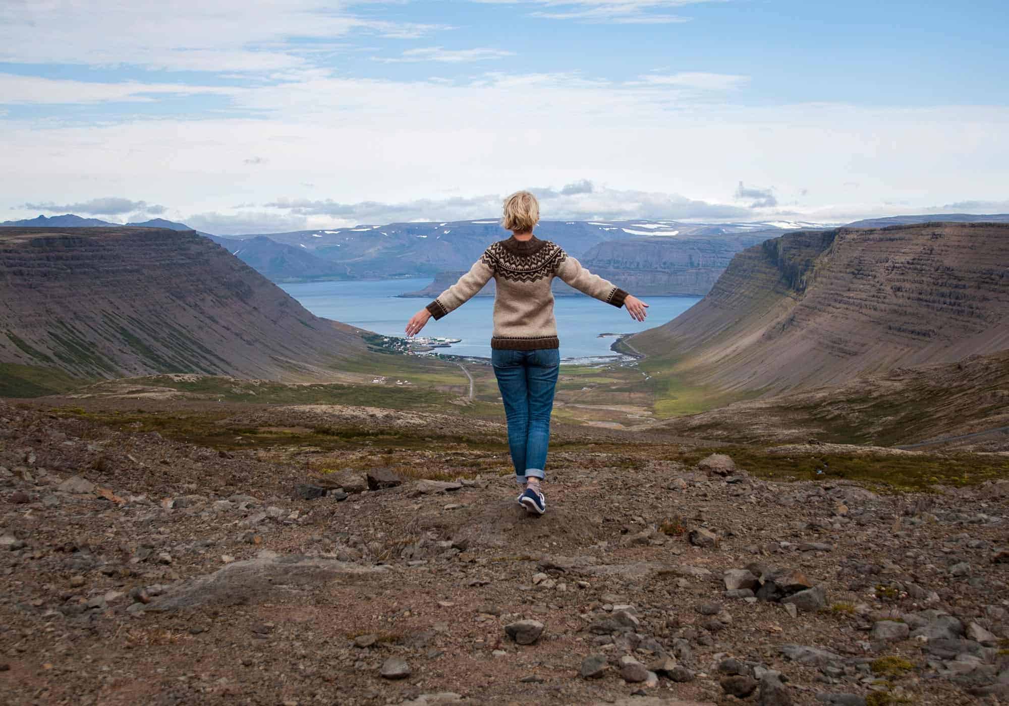 Woman wearing Icelandic lopapeysa sweater stands high above a fjord in west Iceland.
