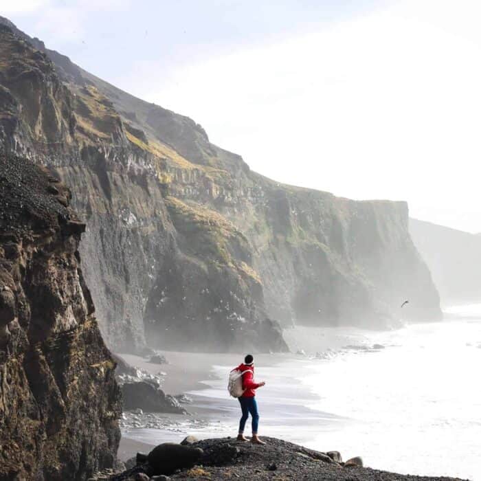 Woman stands on a black sand beach beside basalt cliffs in front of the sea in south Iceland.