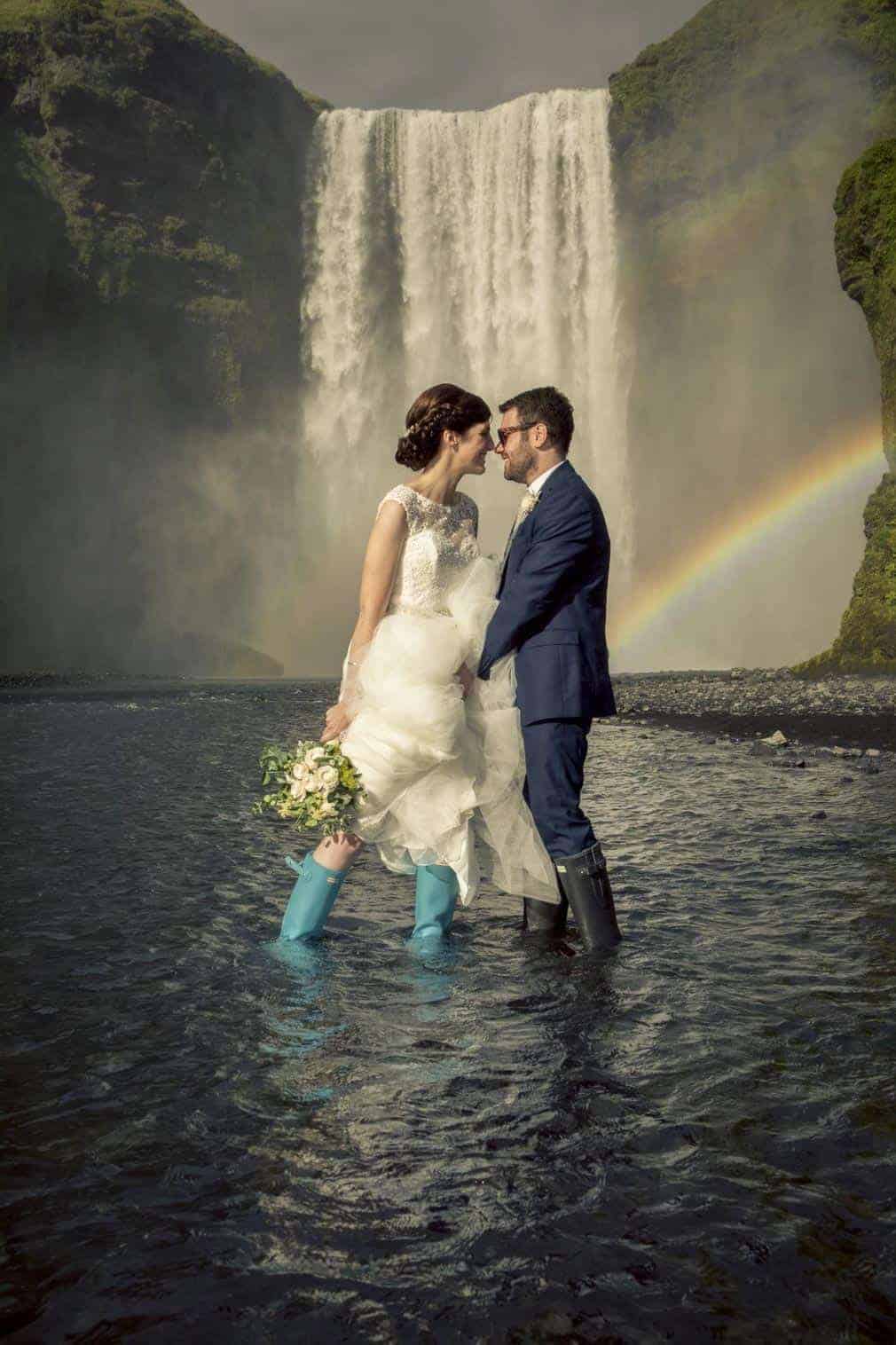 Bride and groom in wedding clothes and rubber boots stand in the river in front of Skógafoss.