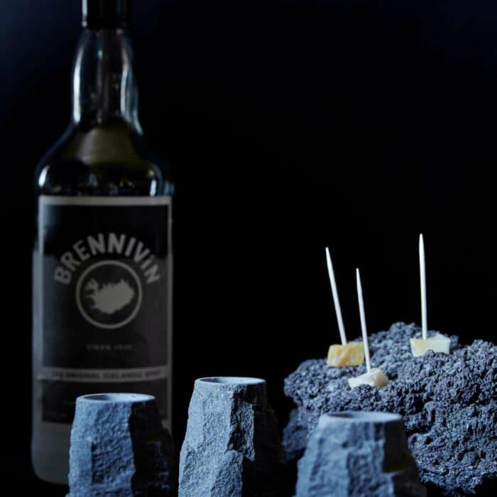 Basalt shot glasses in foreground with Brennivín bottle and hakárl speared with toothpicks.