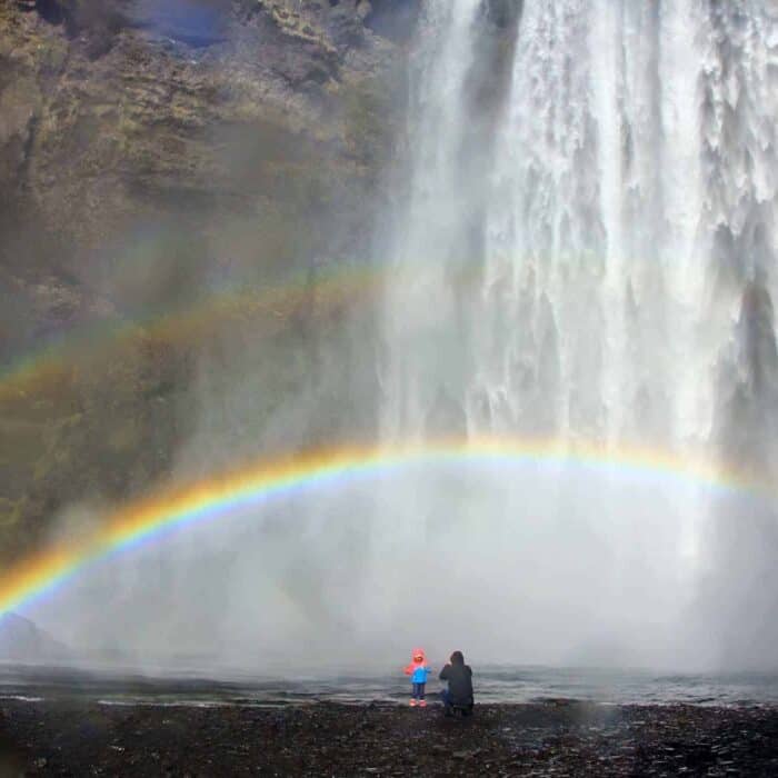 Parent takes photo of child in front of Skógafoss waterfall and rainbows.