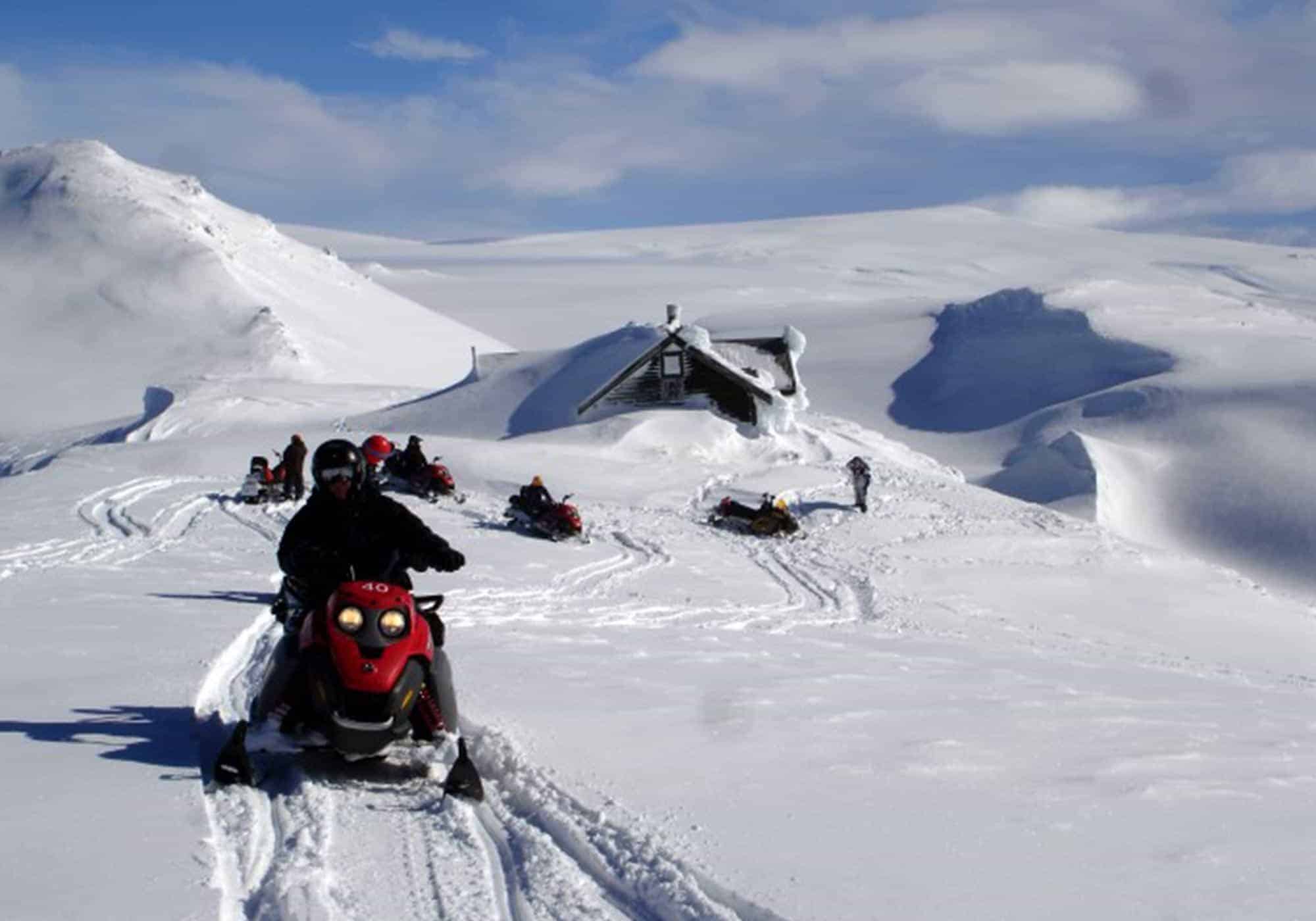 People drive snowmobiles up in the Icelandic highlands beside a snowed-in cabin.
