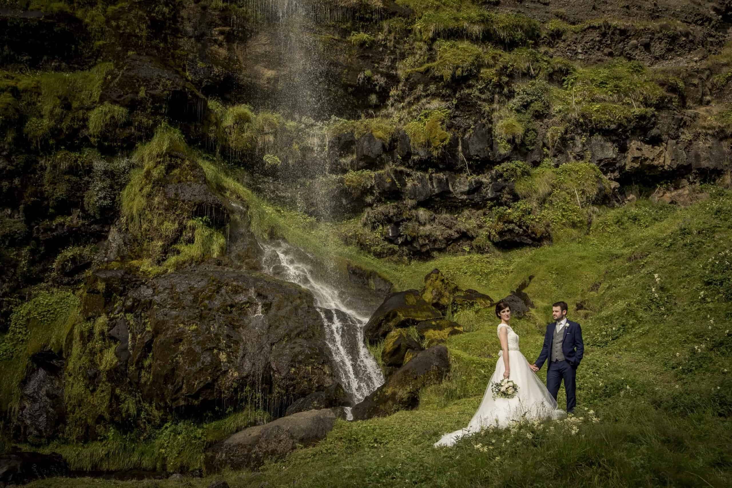 Bride wearing a white dress holds hands with her groom in front of the waterfall Þórðarfoss.