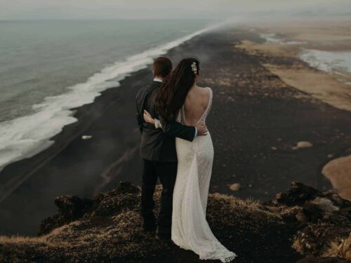 Bride and groom embrace on cliff near Dýrholaey that looks out over a black sand beach.