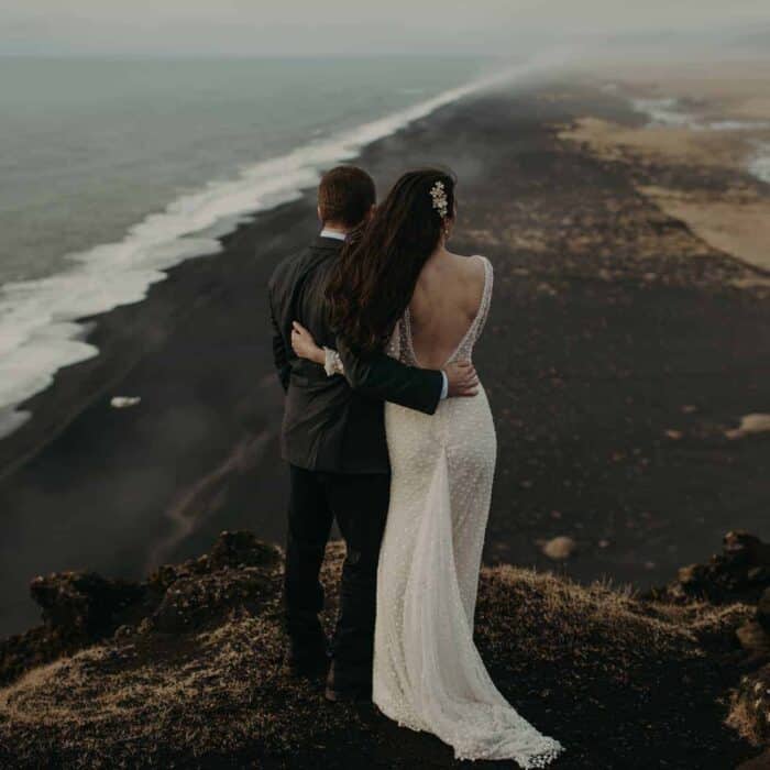 Bride and groom embrace on cliff near Dýrholaey that looks out over a black sand beach.