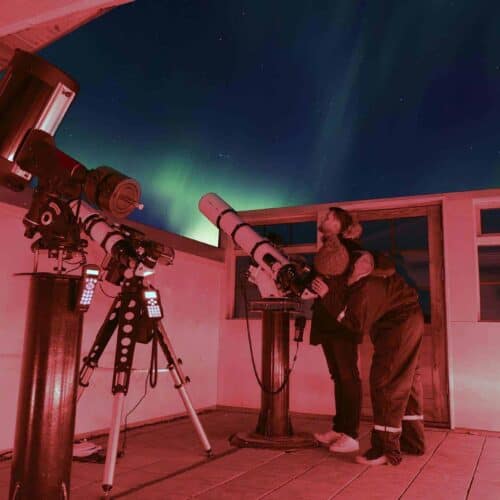Woman peers through a high-powered telescope in the Hotel Rangá Observatory underneath green northern lights.