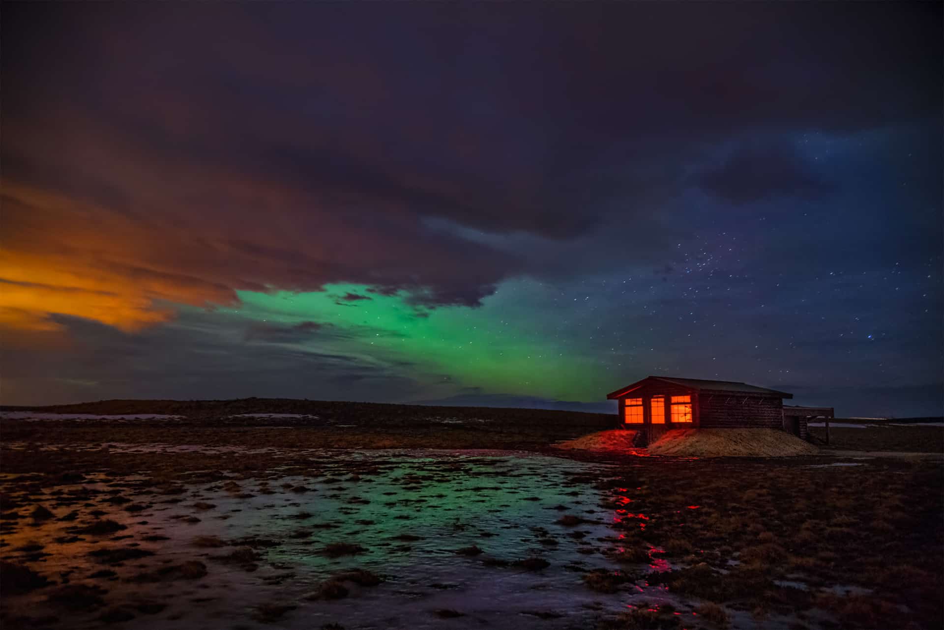 Green and pick northern lights peak from behind clouds over the Hotel Rangá Observatory in south Iceland.