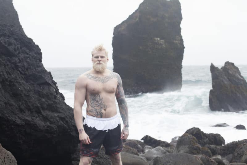 Man wearing Secret of Iceland swim trunks on a black sand beach.