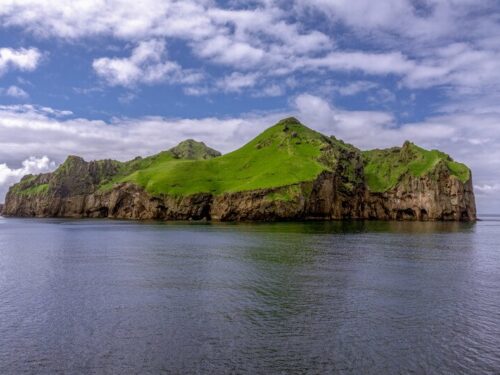 Green grass covers the Westman Islands surrounded with ocean and blue skies overhead.