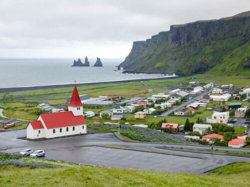 View of Vík village including a small church with a red roof, many houses and a black sand beach with basalt rock formations.