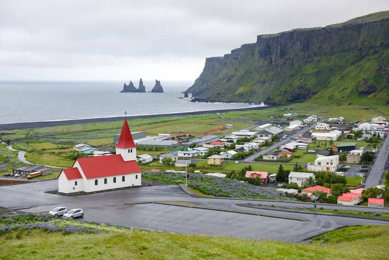 View of Vík village including a small church with a red roof, many houses and a black sand beach with basalt rock formations.