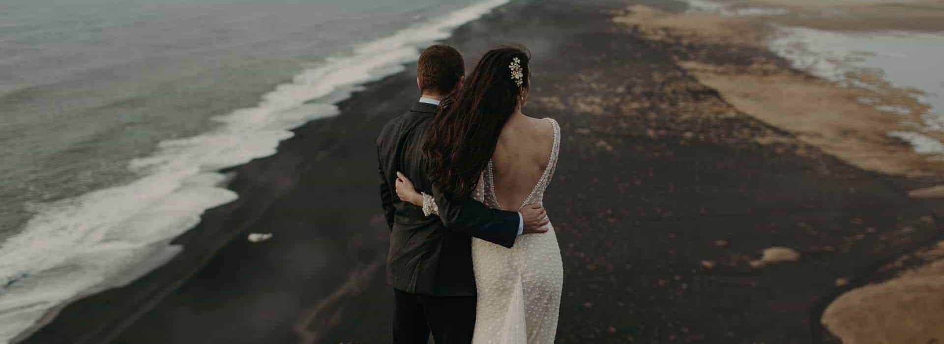 Wedding couple embraces atop the Dýrholaey promontory with views of a black sand beach.