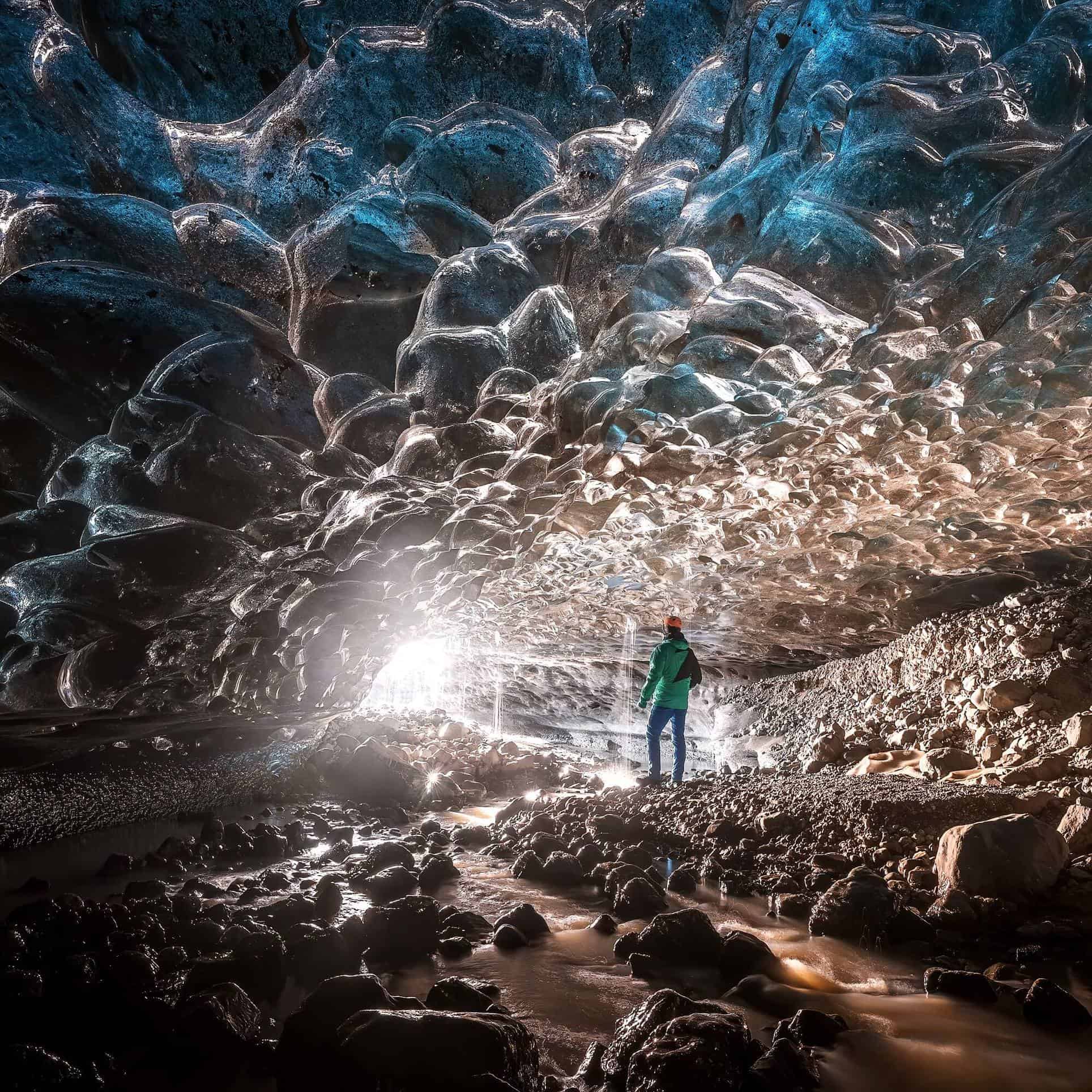 Sun streams into an ice cave where a man stands atop rocky boulders.