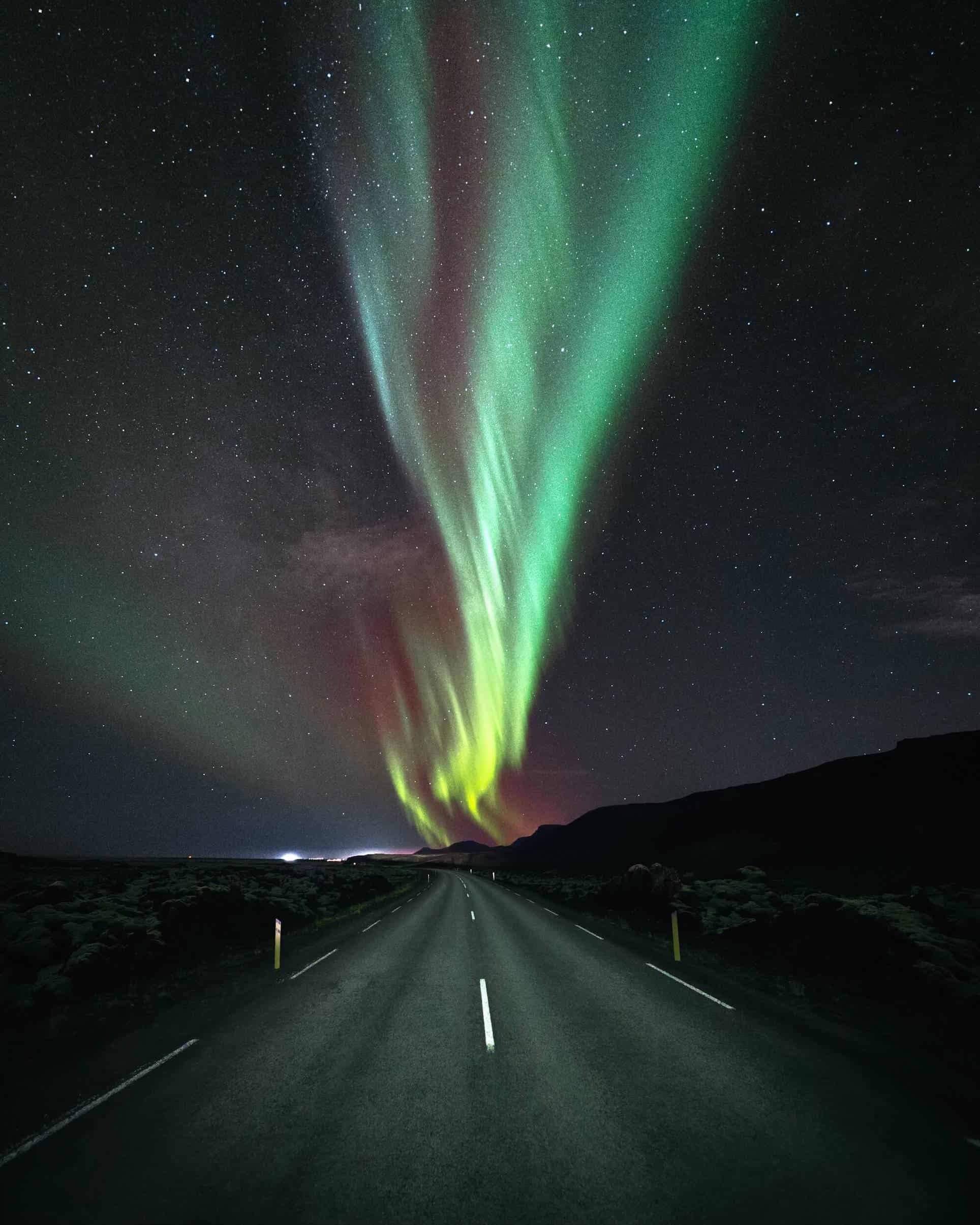 A band of pink and green northern lights shimmer above a paved country road in south Iceland.