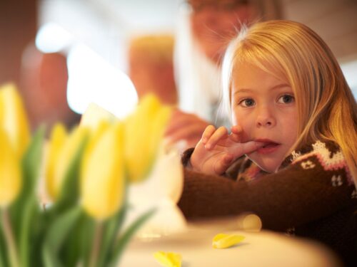 A little blonde girl sitting at a table and sticking her index finger in her mouth with tulips in the foreground.