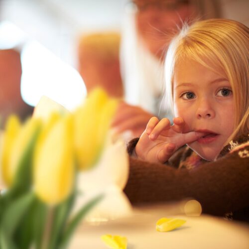 A little blonde girl sitting at a table and sticking her index finger in her mouth with tulips in the foreground.