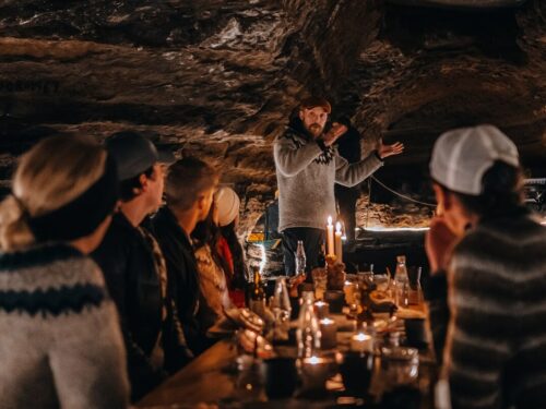 Man stands at the end of a long table filled with candles, food and drink and other guests inside the Caves of Hella.