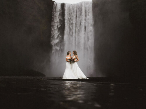 Queer wedding photoshoot of two brides standing in front of Skógafoss waterfall in south Iceland.