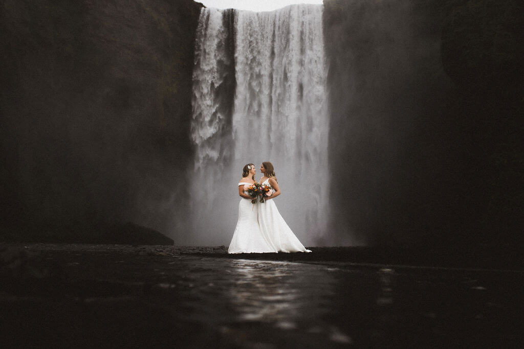 Queer wedding photoshoot of two brides standing in front of Skógafoss waterfall in south Iceland.