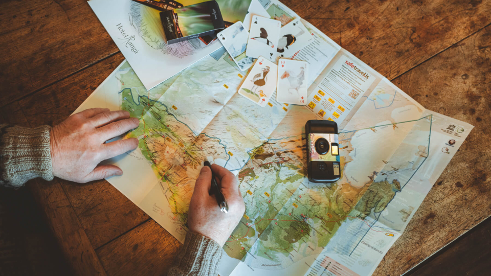 Hands holding a pen draw on a map of Iceland that is beside an analog camera.