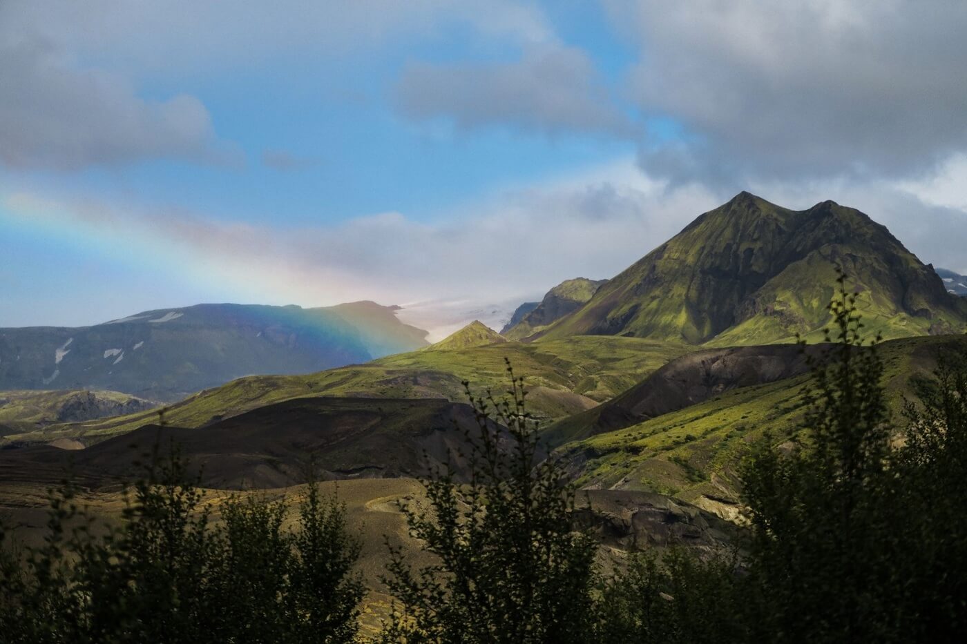 A rainbow in the midst of mossy mountains and small trees underneath cloudy skies in Þórsmörk.