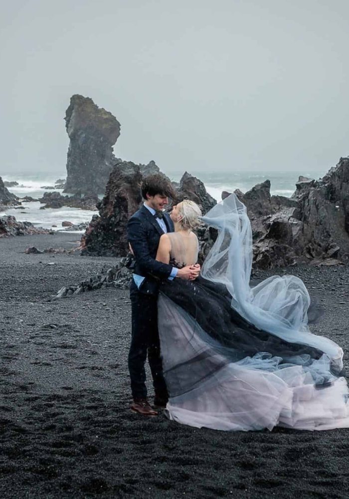 Groom in suit and bride in flowing grey and black wedding dress stand on a black sand beach on a stormy day in south Iceland.